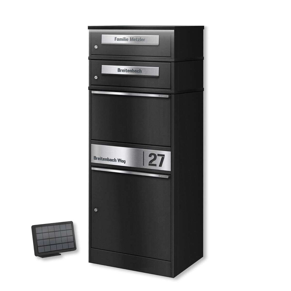 Tall black metal multi-drawer mailbox unit with silver nameplates on each drawer and a large bottom compartment; a small solar panel sits on the floor nearby.