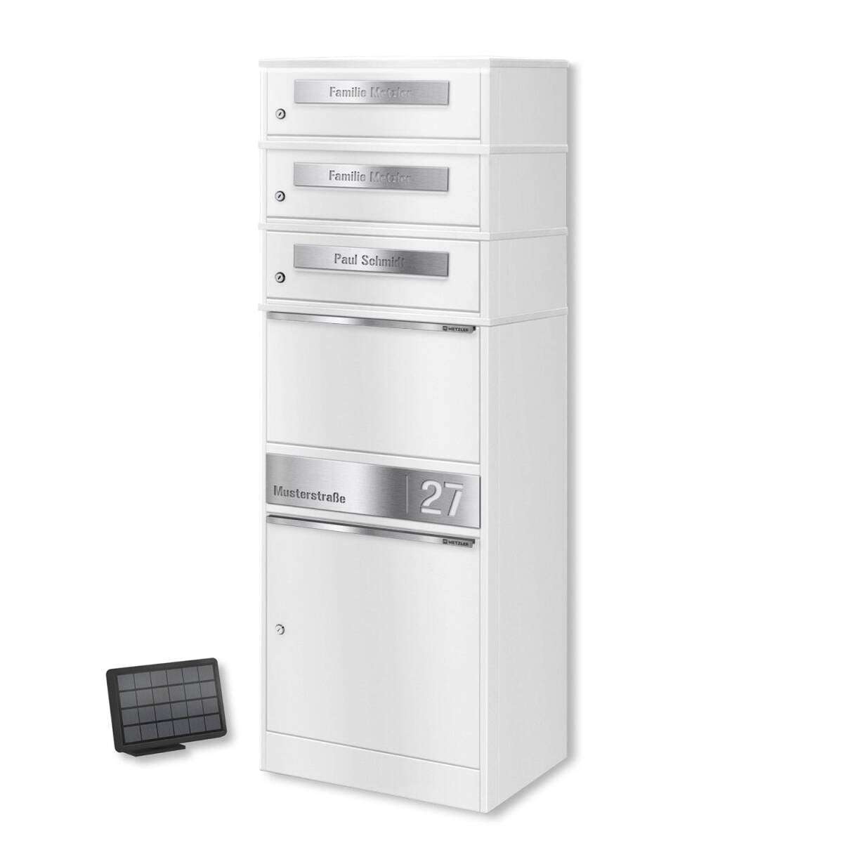 White vertical multi-compartment mailbox with four small drawers and a larger bottom section, featuring metal nameplates on each drawer. A small solar panel sits to the left on the ground.