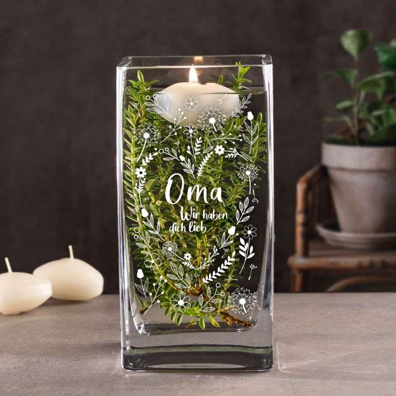 Lit white candle in a glass block holder with greenery inside and white floral designs, text reads 'Oma Wir haben dich lieb'.