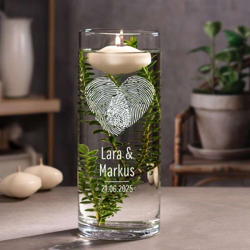 Tall glass hurricane with floating candle and green foliage, wedding name card reading 'Lara & Markus' and date '21.06.2025' inside a heart motif, decorative keepsake.