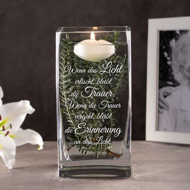 Glass memorial candle in a rectangular crystal vase with green sprigs and a lit white candle; German inscription about light and memory.